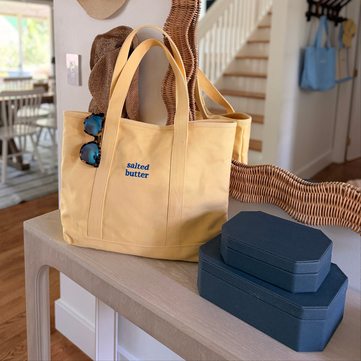 A yellow canvas tote bag with blue 'salted butter' text sits on a light wood console table in a home entryway. The bag has sunglasses hanging from the handle and is styled with a blue box on a matching blue stool nearby, with a wavy mirror and straw hats in the background.