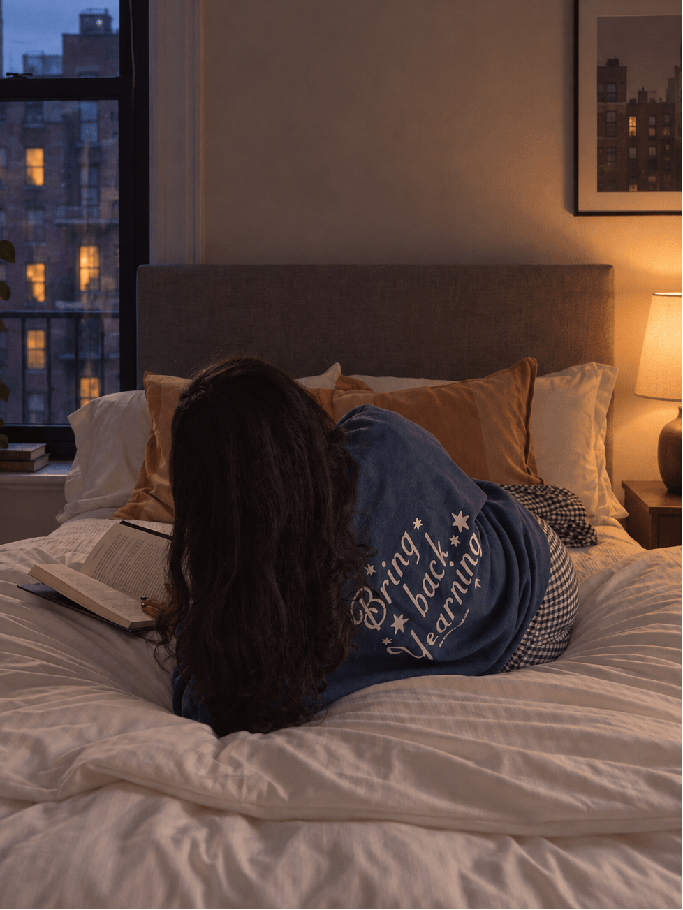A person lies on a bed in a warmly lit bedroom, wearing a blue sweatshirt with white text that reads 'Brigade, learn.' A window with a city skyline at dusk is visible on the left, and a lamp glows on the nightstand.