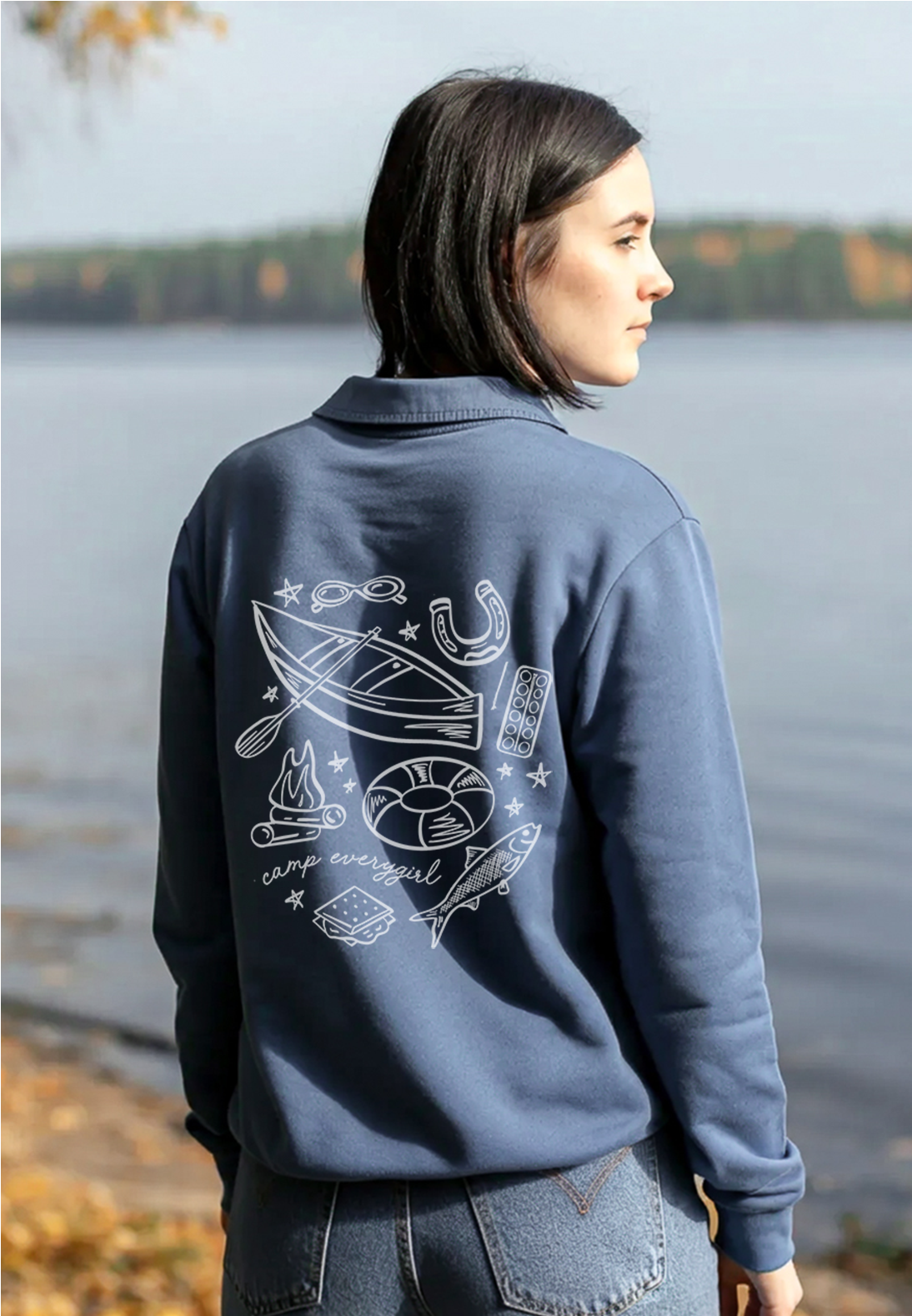 Person wearing a blue jacket with a graphic design on the back, standing by a lake.