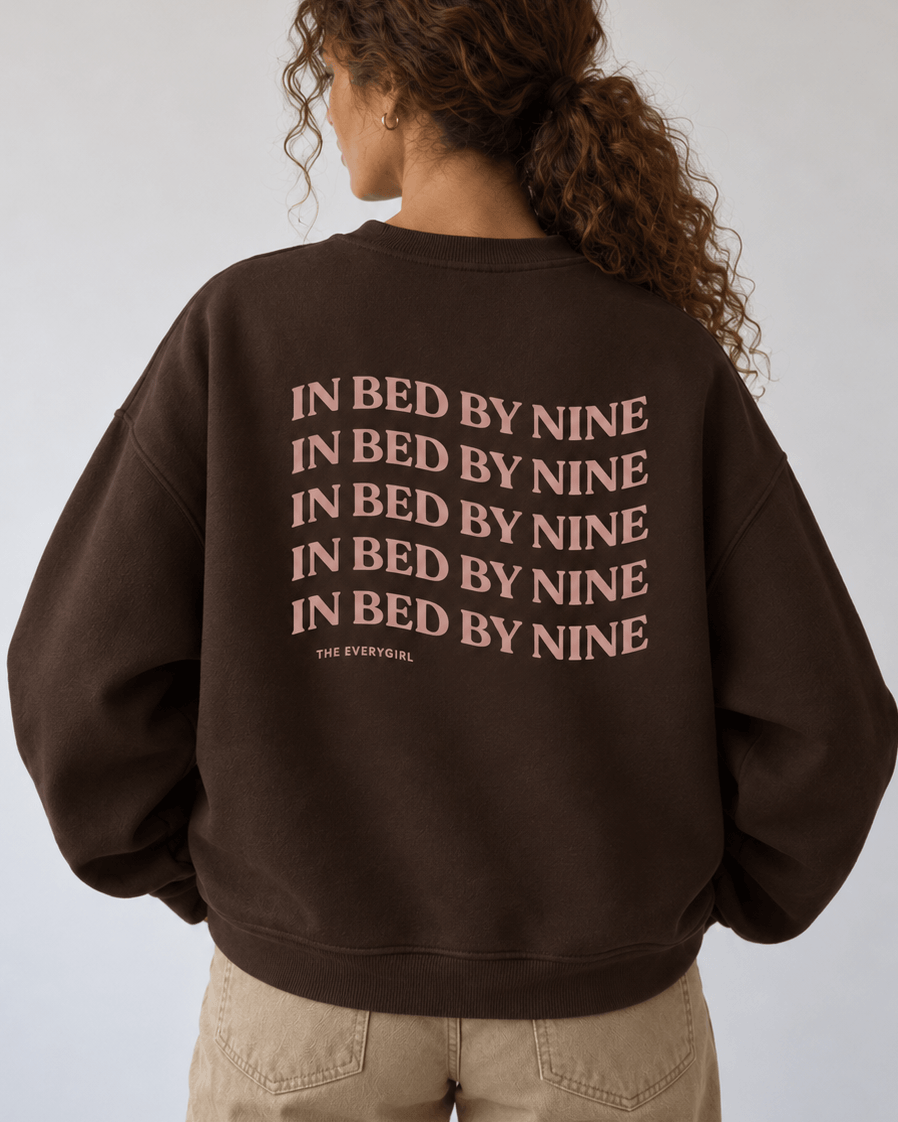 A person shown from the back wearing a dark brown sweatshirt with the phrase 'IN BED BY NINE' repeated in a muted pinkish-beige font across the back, paired with light beige pants against a plain light gray studio background.