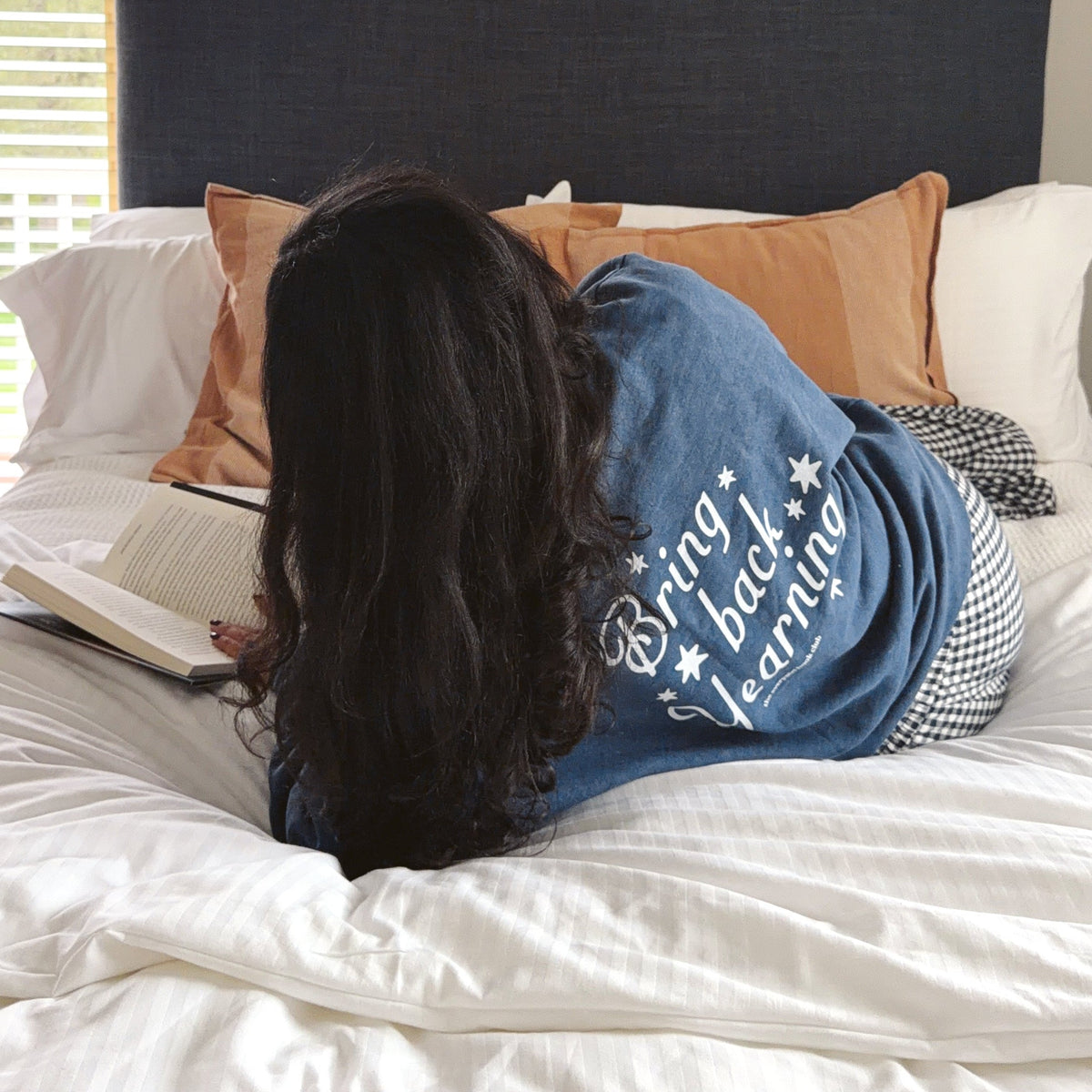 A woman lies on a bed reading an open book, viewed from behind. She is wearing a blue denim-style shirt with white text that reads 'Bringing bad learning'.