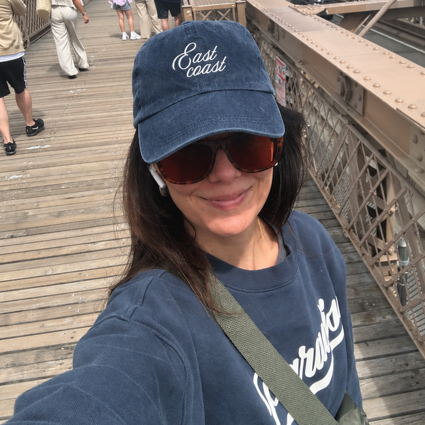 Person wearing a blue cap and sunglasses on the Brooklyn Bridge with a city skyline in the background.